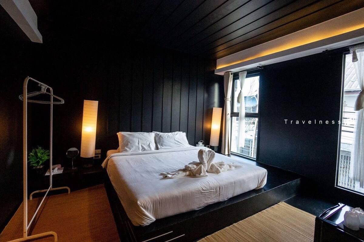 A modern hotel room with a large bed, bedside lamps, a view of the city, and a black wall with the word "Travelness".