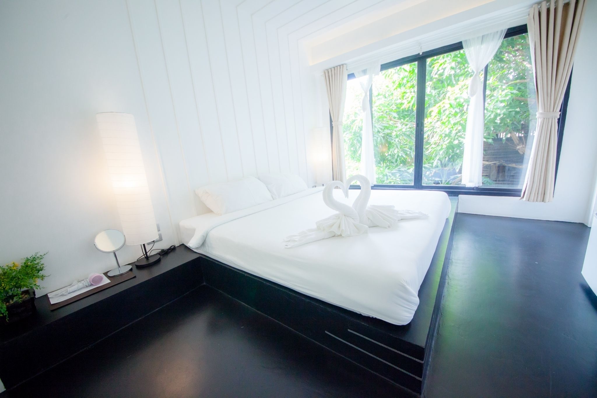 Una cama bien hecha con ropa de cama blanca y una disposición de toallas en forma de corazón. Hay una mesita de noche con una lámpara y un pequeño espejo. La habitación tiene un ventanal con cortinas y vistas a zonas verdes.