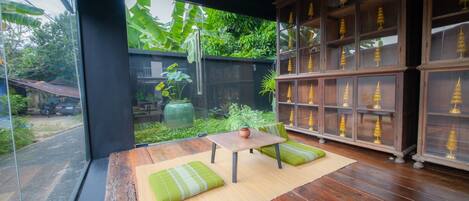 A room with a wooden floor, a table with a vase, green cushions, and a view of a garden with trees and a large window.