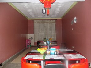 A dining area with a table set for four, a pink chair, a red chair, and a small table with a basket of fruit. - Hotel B Plus (Mbarara)