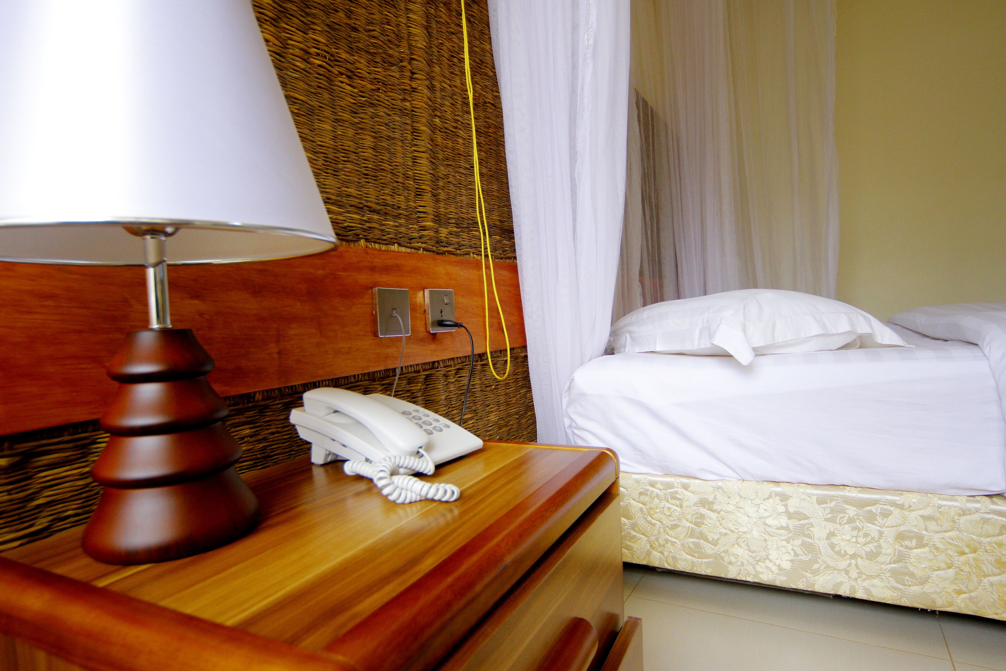 Una habitación de hotel con una mesita de noche de madera, una lámpara blanca, un teléfono y una cama con ropa blanca.