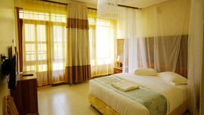 A bedroom with a bed, curtains, a bedside table with a lamp, and a television on a stand. - Kalya Courts Hotel (Fort Portal)