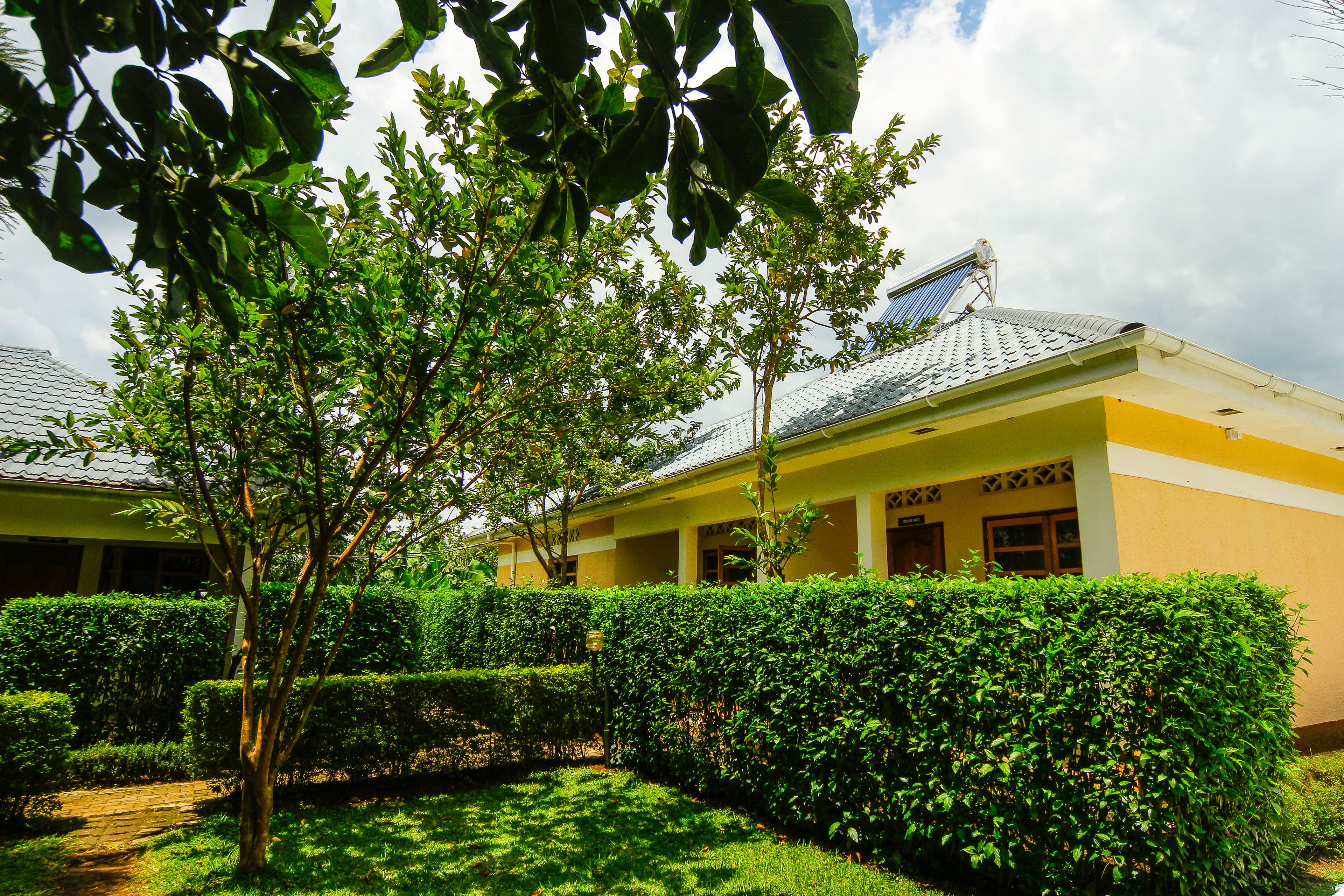 A building with a yellow facade, a covered walkway, and a well-maintained garden with hedges and trees.