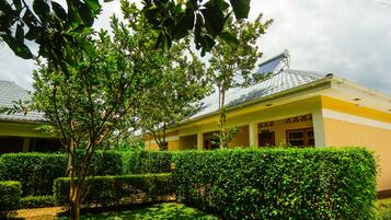 A building with a yellow facade, a covered walkway, and a well-maintained garden with hedges and trees.