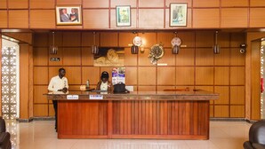 A reception desk with two staff members, wooden paneling, framed pictures, and a lion statue. - Kalya Courts Hotel (Fort Portal)
