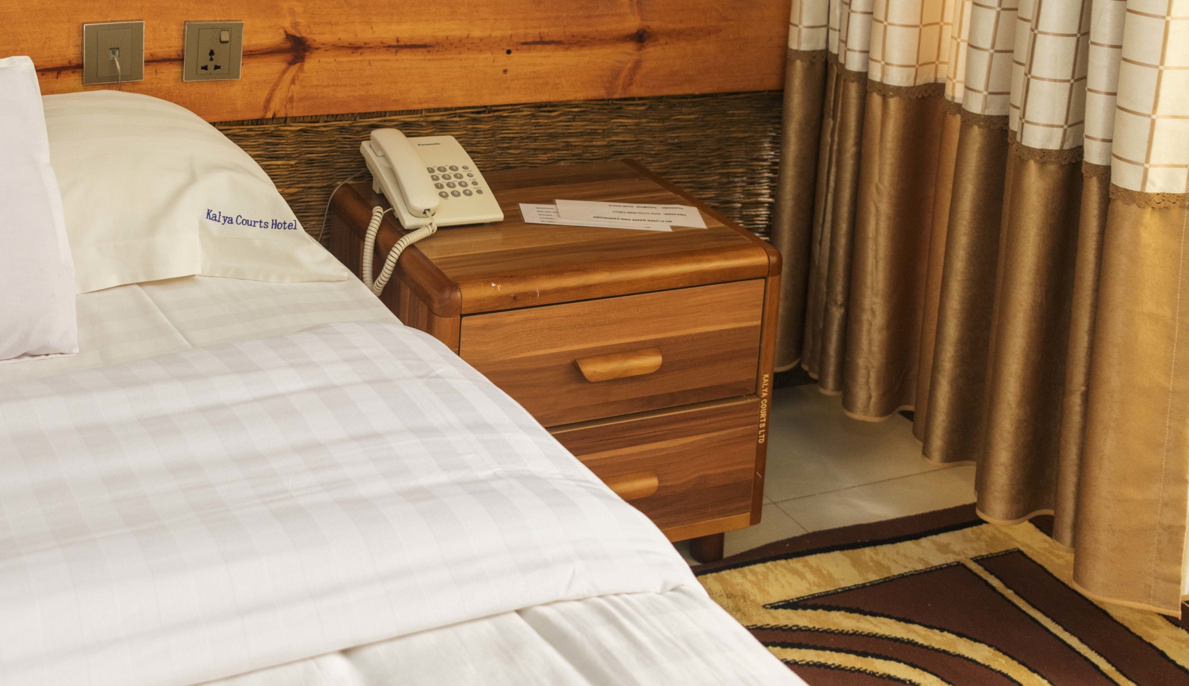 A hotel room with a bed, a wooden nightstand with a phone, and a wall socket.