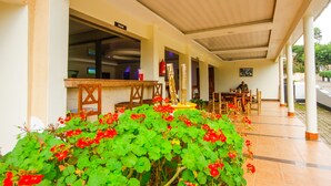 A covered outdoor area with a bar, seating, and a garden with red flowers. - Kalya Courts Hotel (Fort Portal)