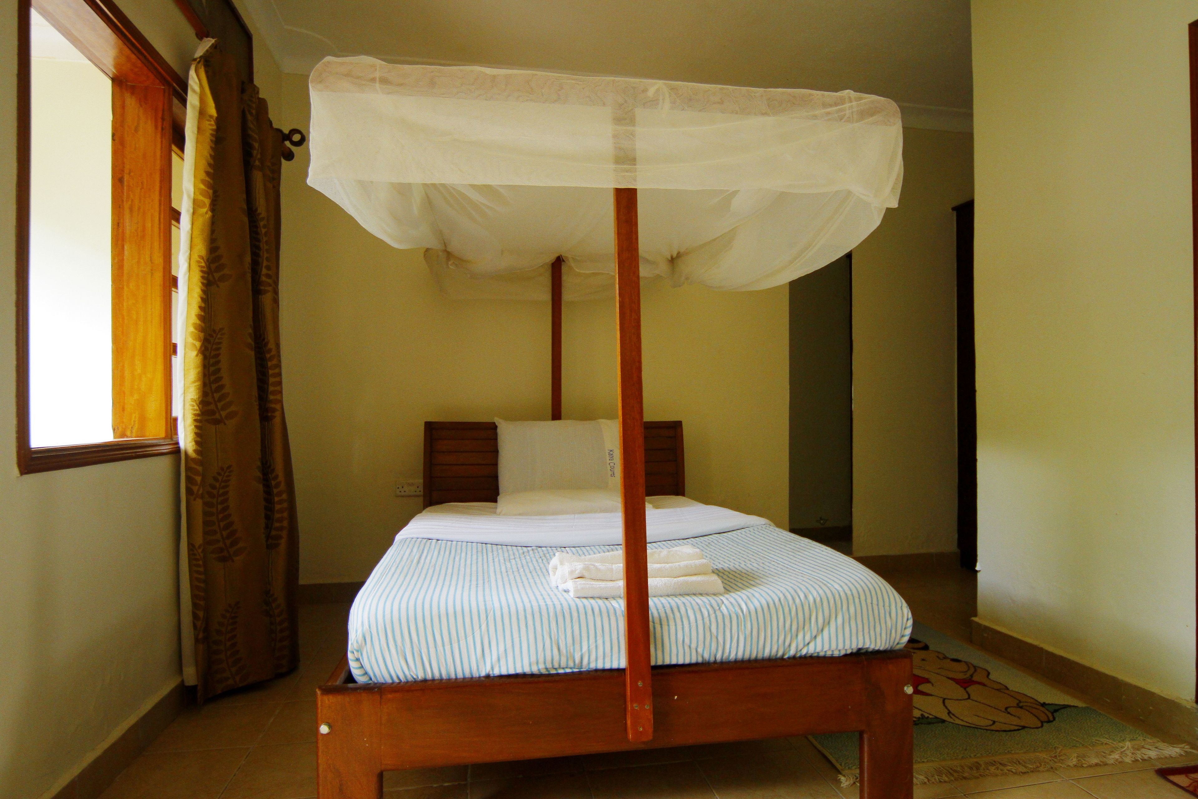 A single bed with a wooden frame and a canopy, a window with curtains, and a cartoon character rug on the floor.