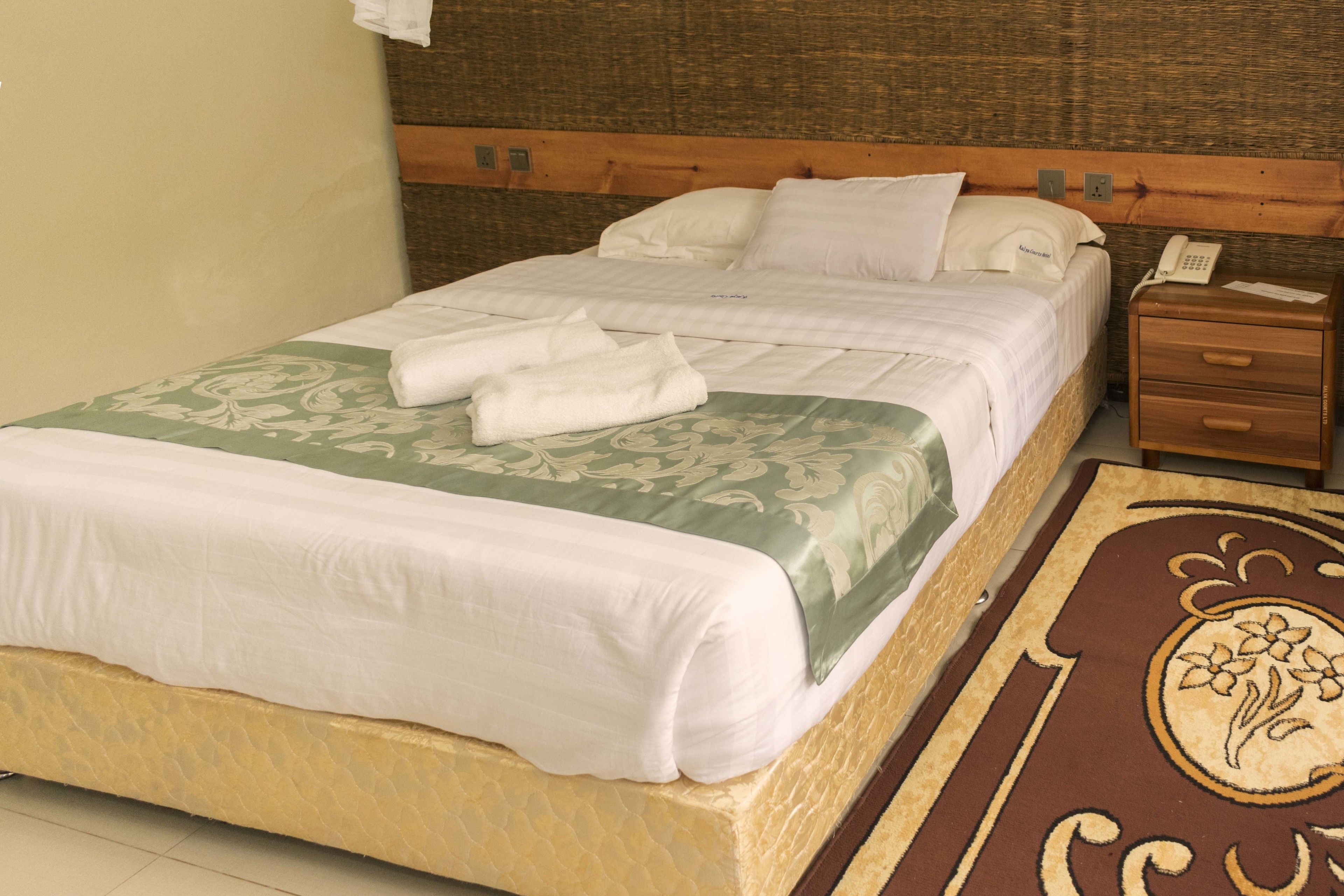 A neatly made bed with a green and white bedspread, a wooden headboard, a bedside table with a telephone, and a decorative rug.