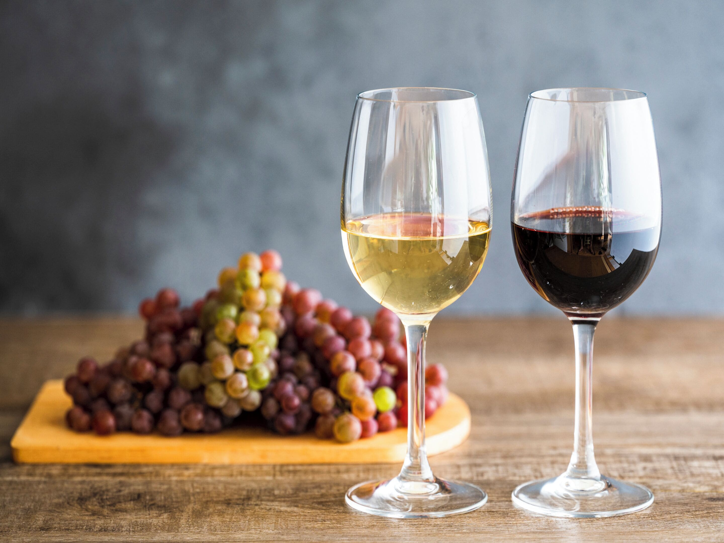 Two wine glasses, one with white wine and the other with red wine, on a wooden surface with a pile of grapes in the background.