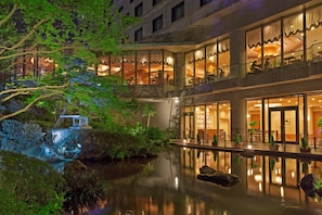 Un hotel con un patio tranquilo que cuenta con un estanque reflectante, abundante vegetación y un edificio de estilo tradicional con varios balcones e iluminación cálida.