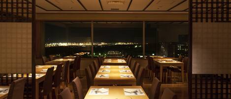 Un restaurant doté de nombreuses tables et chaises, avec de grandes fenêtres offrant une vue sur la ville, en particulier la nuit.