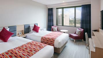 Habitación de hotel con dos camas, un escritorio, una silla, una mesita y un ventanal con vistas a la ciudad.