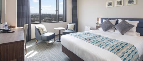 A hotel room with a large bed, a desk, a chair, a small round table, and a view of the city through large windows.