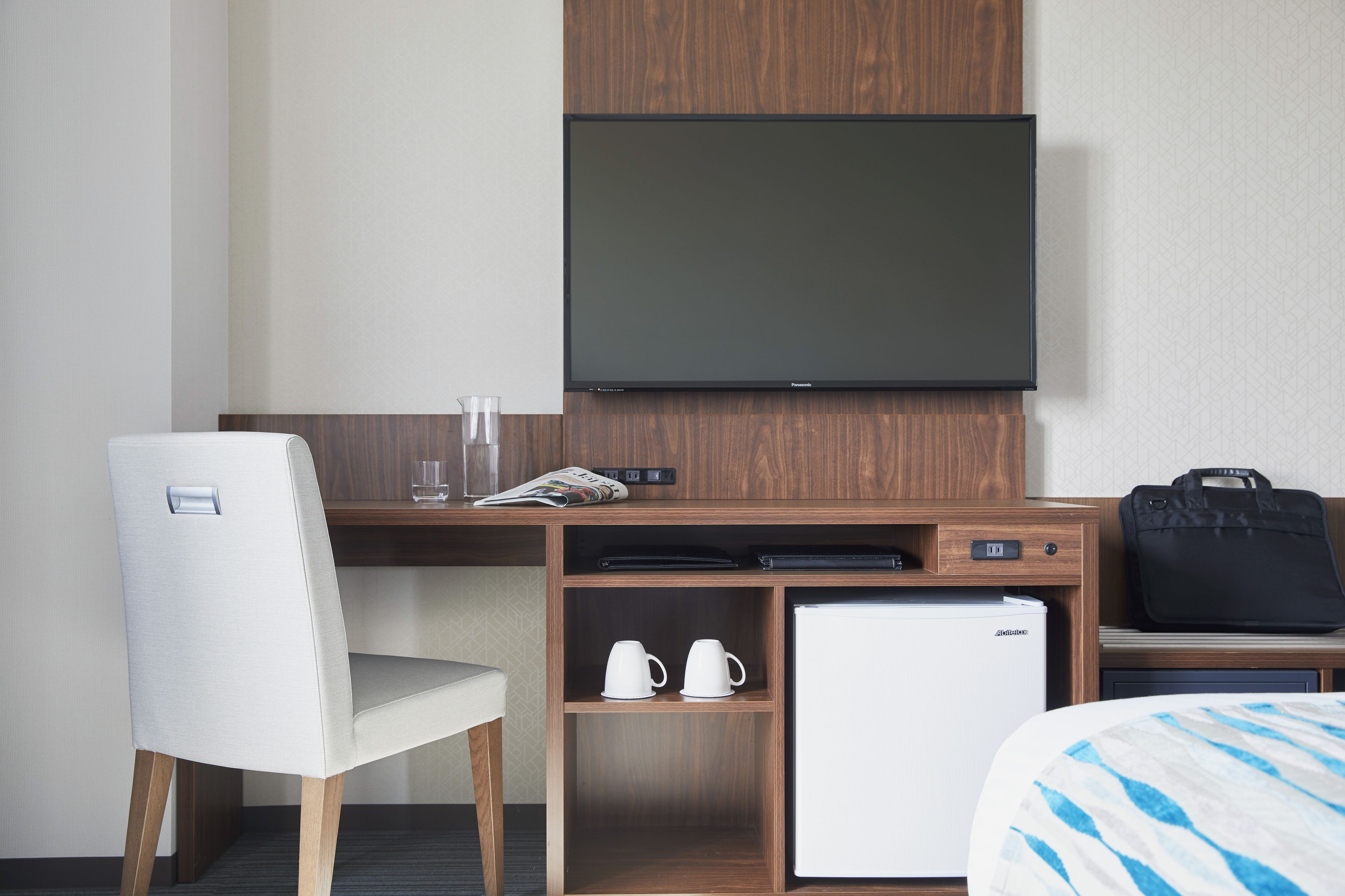 Une chambre d’hôtel comprenant un lit, un bureau avec une télévision, un réfrigérateur, une chaise et un petit meuble avec des tasses.