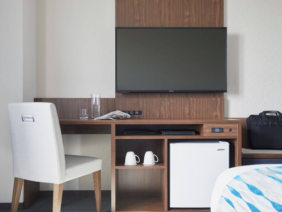 Une chambre d’hôtel comprenant un lit, un bureau avec une télévision, un réfrigérateur, une chaise et un petit meuble avec des tasses.