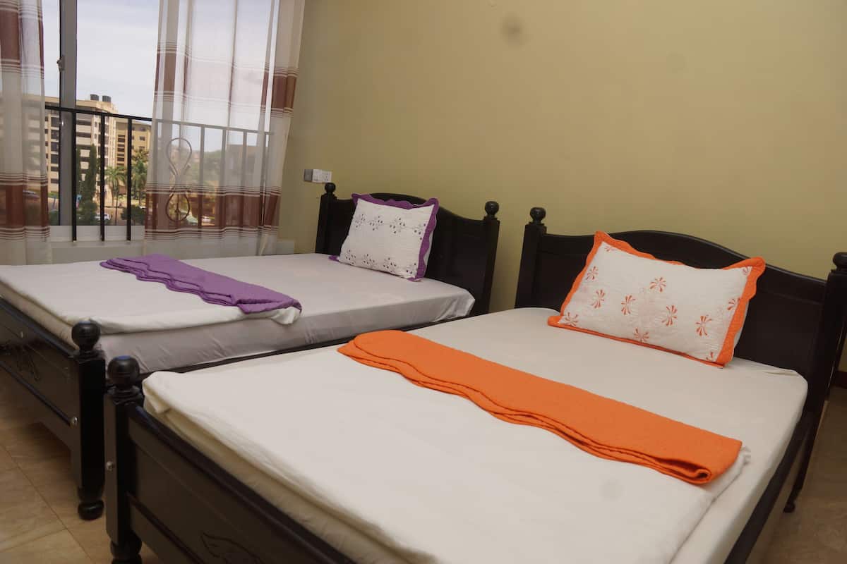 Two single beds with different colored pillows and blankets, a window with curtains, and a view of buildings outside.