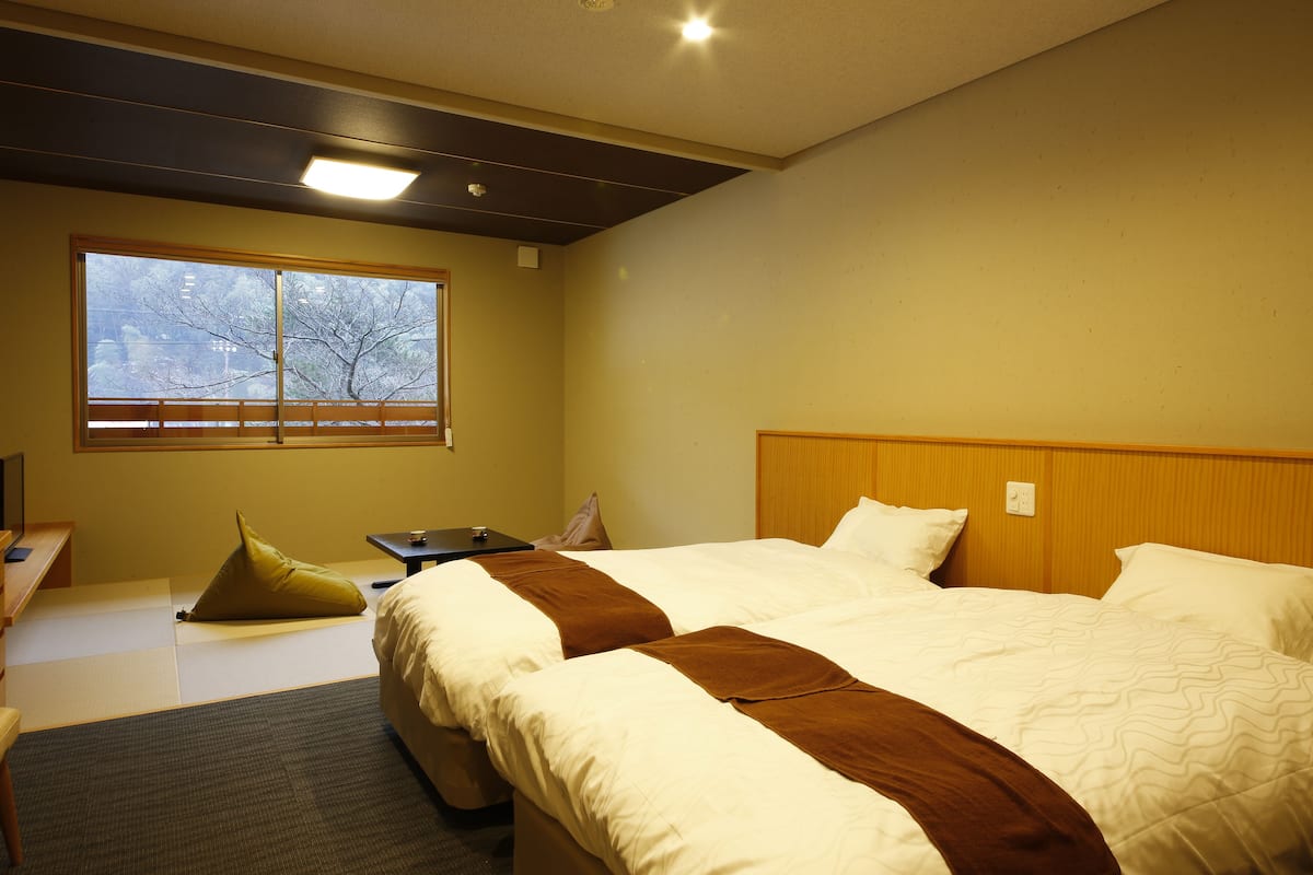 Quarto de hotel com duas camas, uma escrivaninha, uma cadeira, uma janela e vista para árvores lá fora.