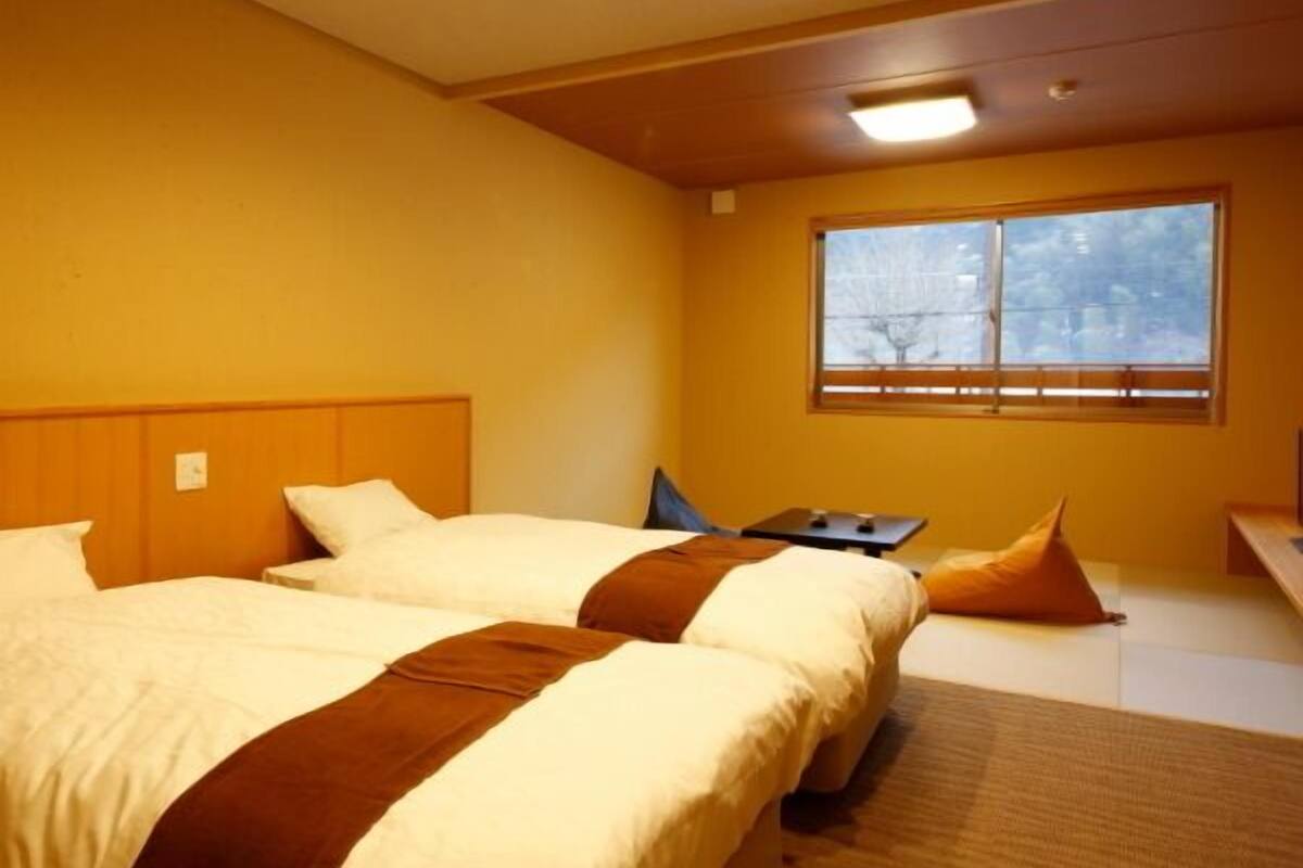 Quarto de hotel com duas camas, uma mesinha, janela com vista e luminária no teto.