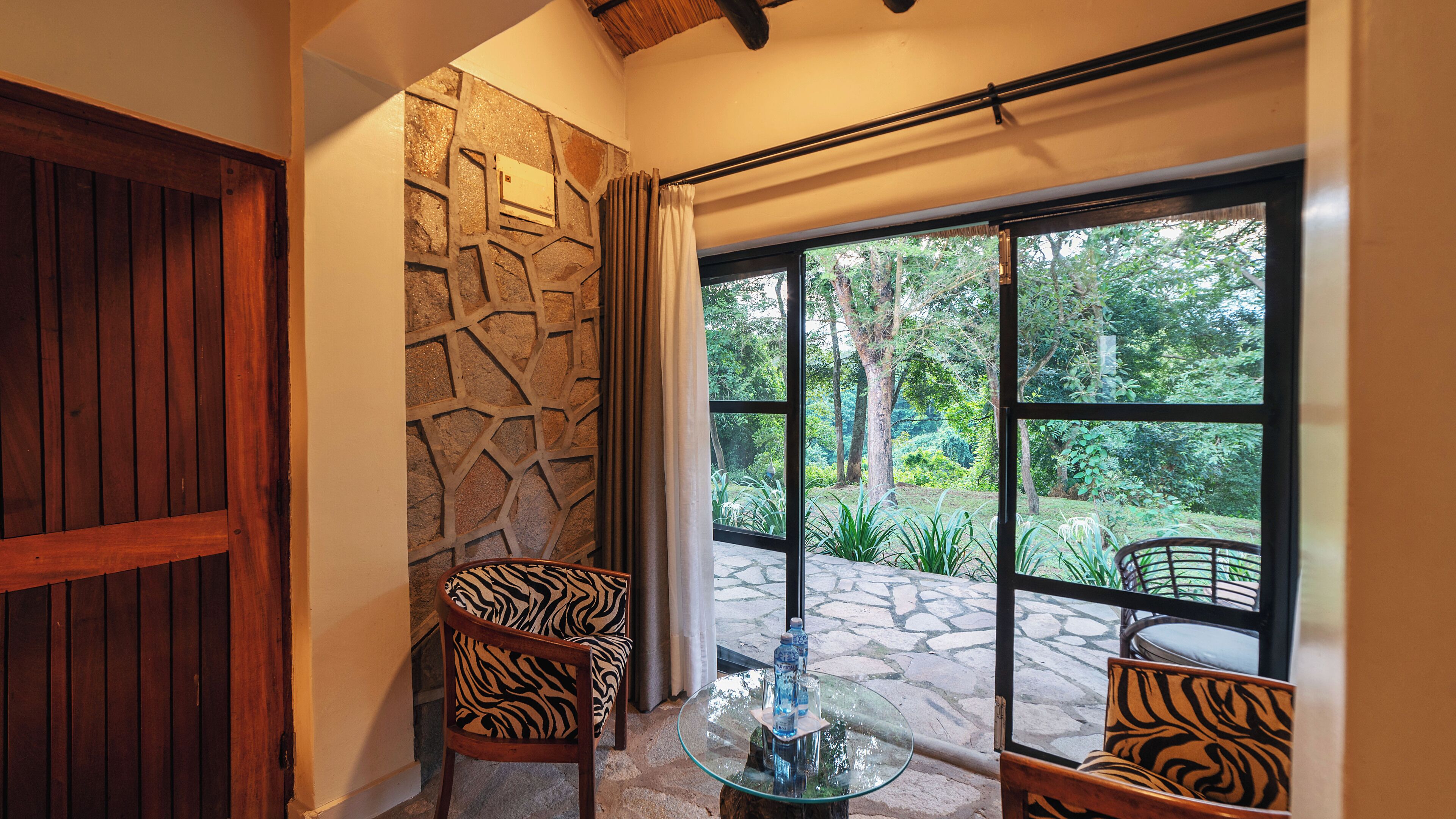 A room with a stone wall, a wooden door, and a large window with a view of greenery. There are two zebra-patterned chairs and a glass table with a bottle on it.
