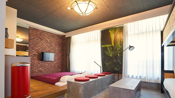 A modern room with a brick accent wall, a flat-screen TV, a large window with sheer curtains, a red trash bin, and a concrete bench with red cushions. - Hotel Material (Kyoto)