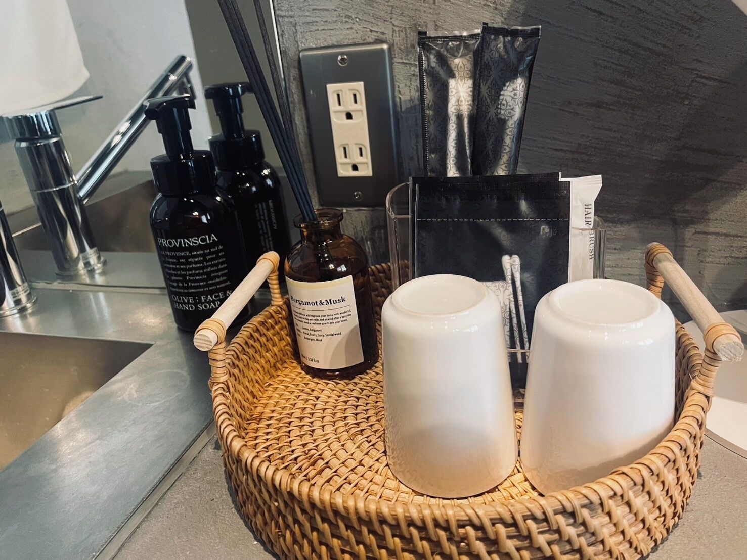 A bathroom counter with toiletries, a bottle of Bergamot & Musk, and two white cups.