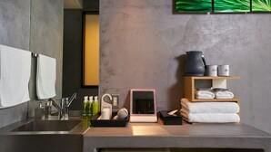 A modern bathroom with a large mirror, a sink with faucets, a towel rack, a shelf with toiletries, and a framed picture. - Hotel Material (Kyoto)