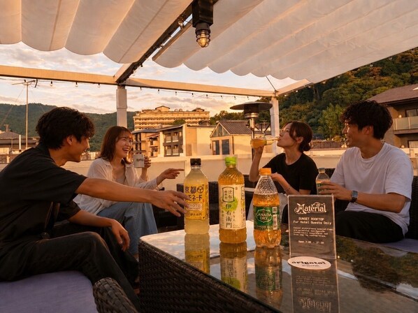 A group of people sitting at an outdoor table with beverages, under a canopy, with a view of buildings and hills in the background. - Hotel Material (Kyoto)
