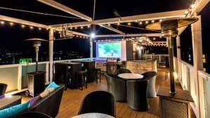 An outdoor patio with wicker furniture, a bar area, and a large screen displaying a golf game. - Hotel Material (Kyoto)
