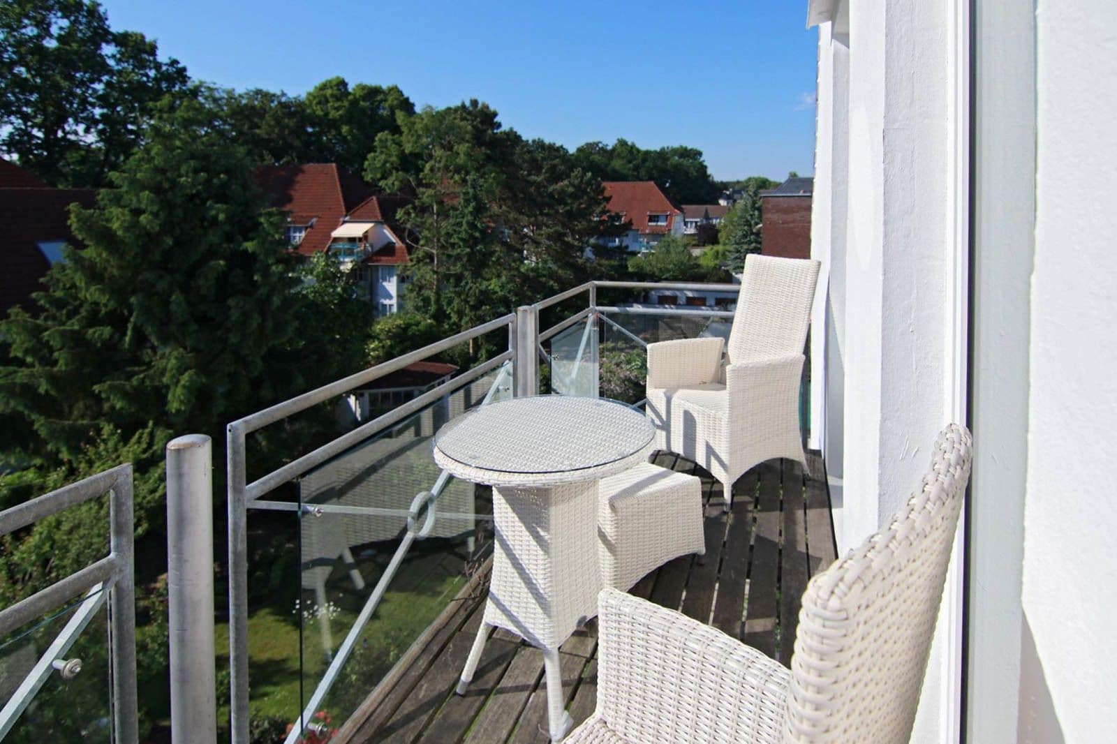 Ein Balkon mit Korbmöbeln und einem Glastisch, mit Blick auf eine Wohngegend mit Häusern und Bäumen.