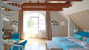 Une chambre d’hôtel moderne avec un plafond en bois, un lit recouvert d’une courtepointe bleue et blanche, un bureau avec une chaise, et une vue sur l’extérieur grâce à une grande fenêtre.