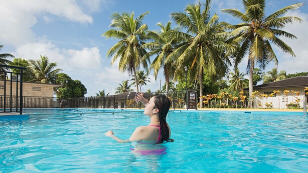 A swimming pool with clear blue water, surrounded by palm trees and a fence. - Pacific Palm Resort (Saipan)