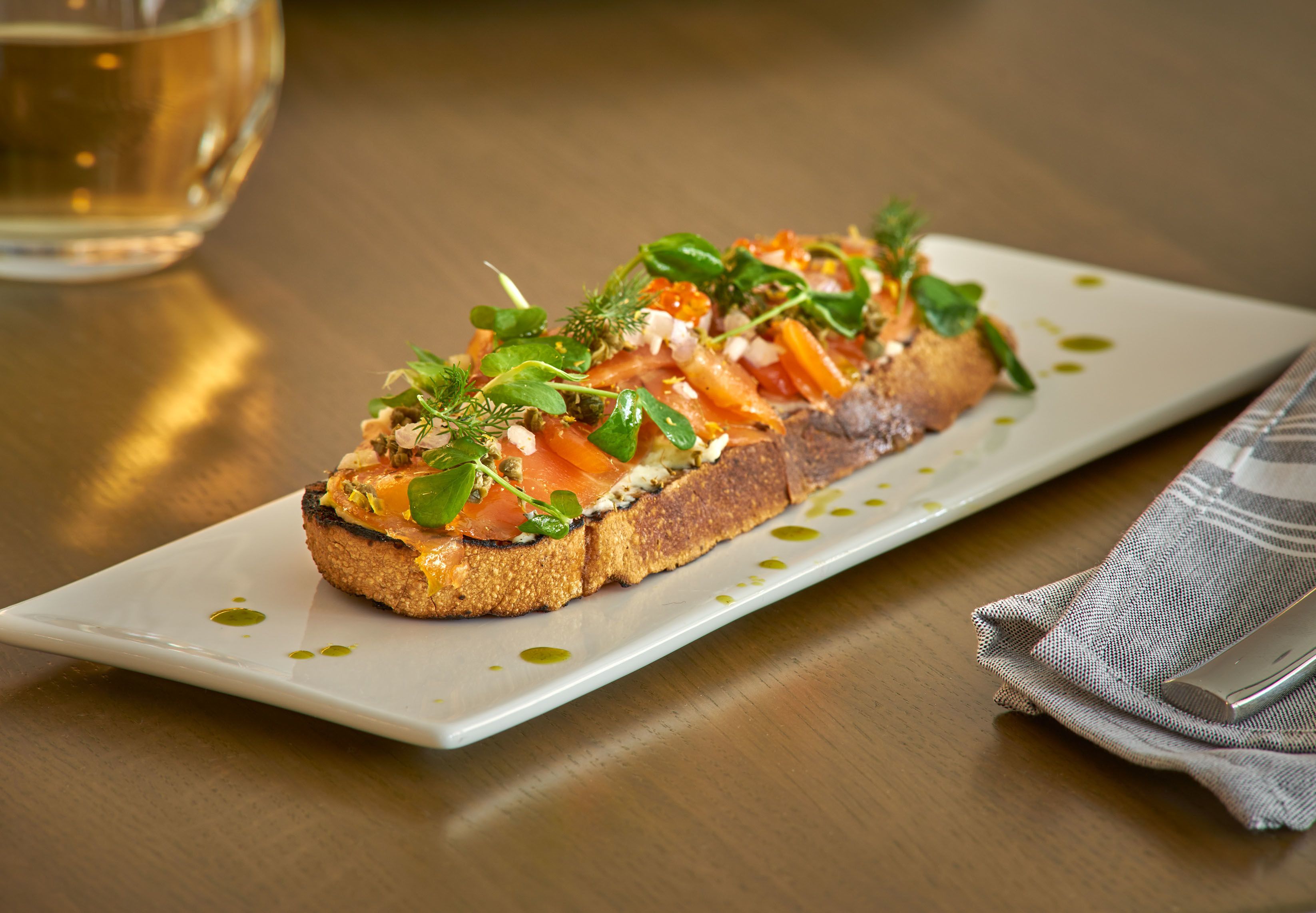 Plat rectangular blanc amb una làmina de pa torrat cobert de salmó, verdures i un raig d'oli d'oliva.