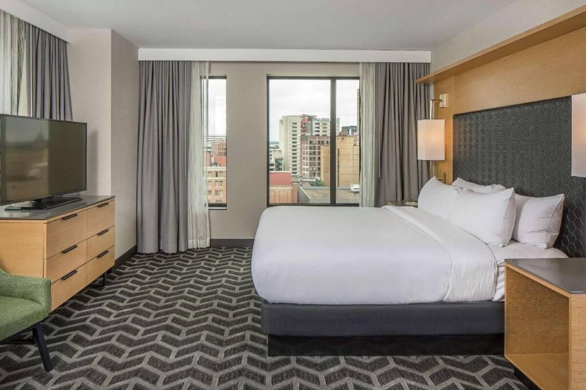 A hotel room with a bed, a TV, a desk, a chair, and a view of the city.