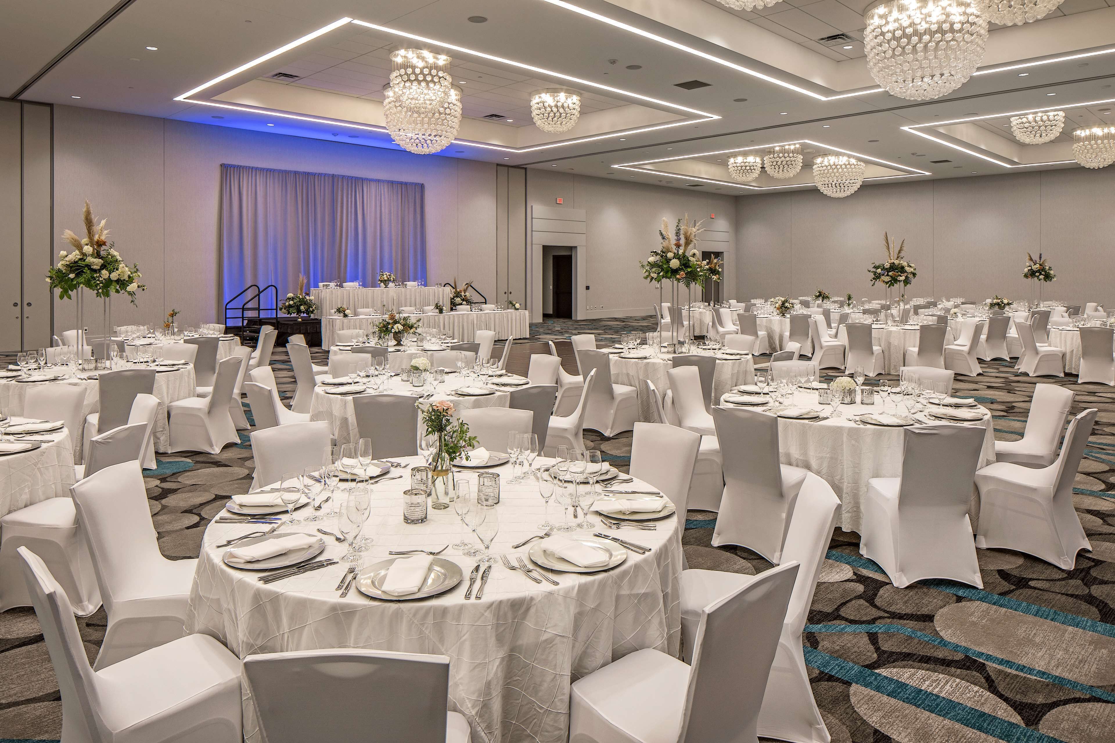 A banquet hall with round tables, white chairs, and chandeliers.