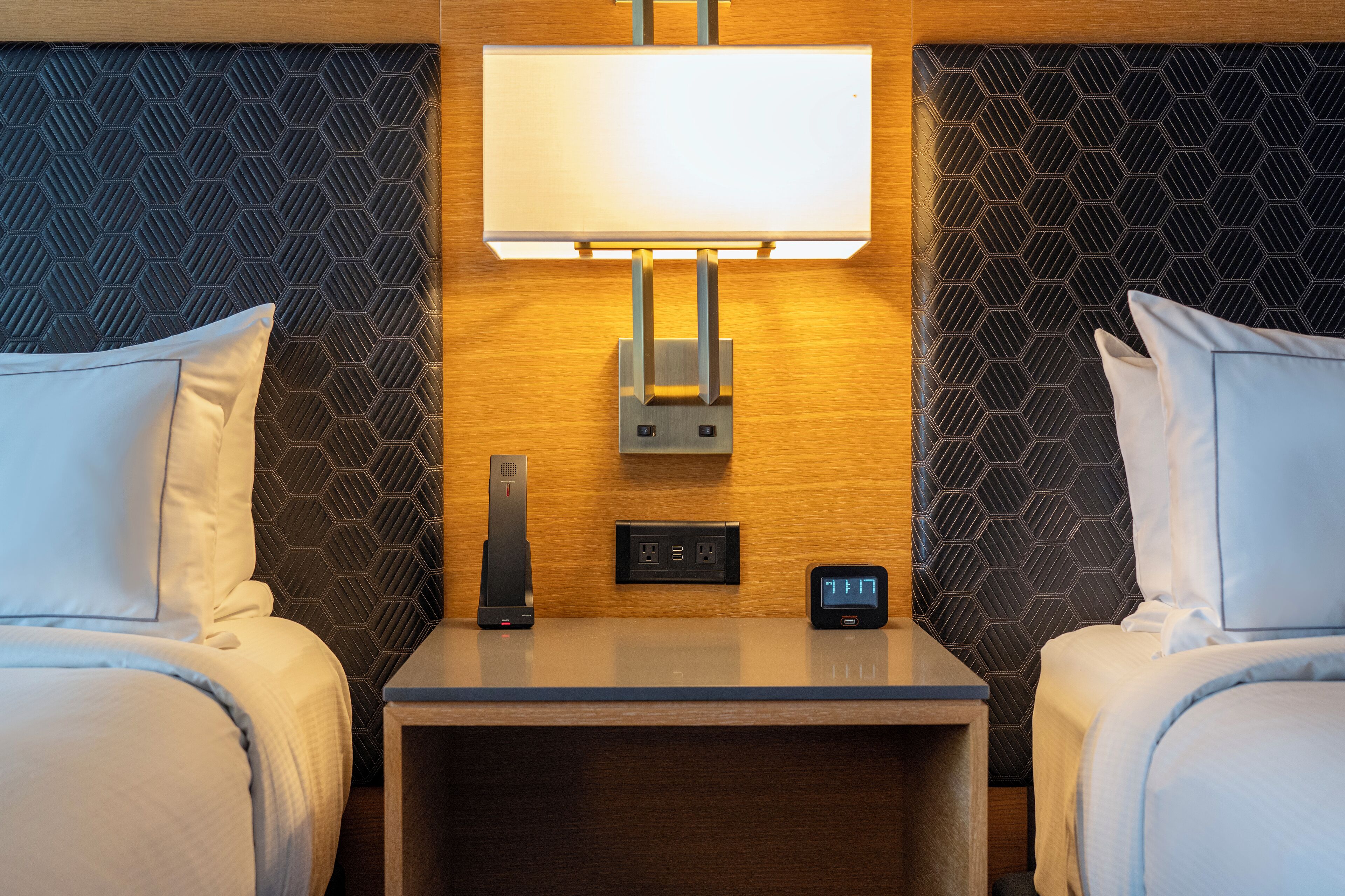 Habitación de hotel con dos camas, una mesita de noche con lámpara, un reloj digital y un enchufe.