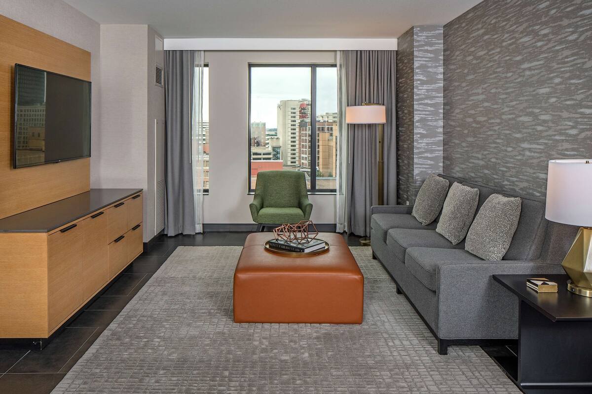 A modern hotel room with a large window, a green armchair, a grey sofa with patterned cushions, a wooden cabinet, and a flat-screen TV mounted on the wall.