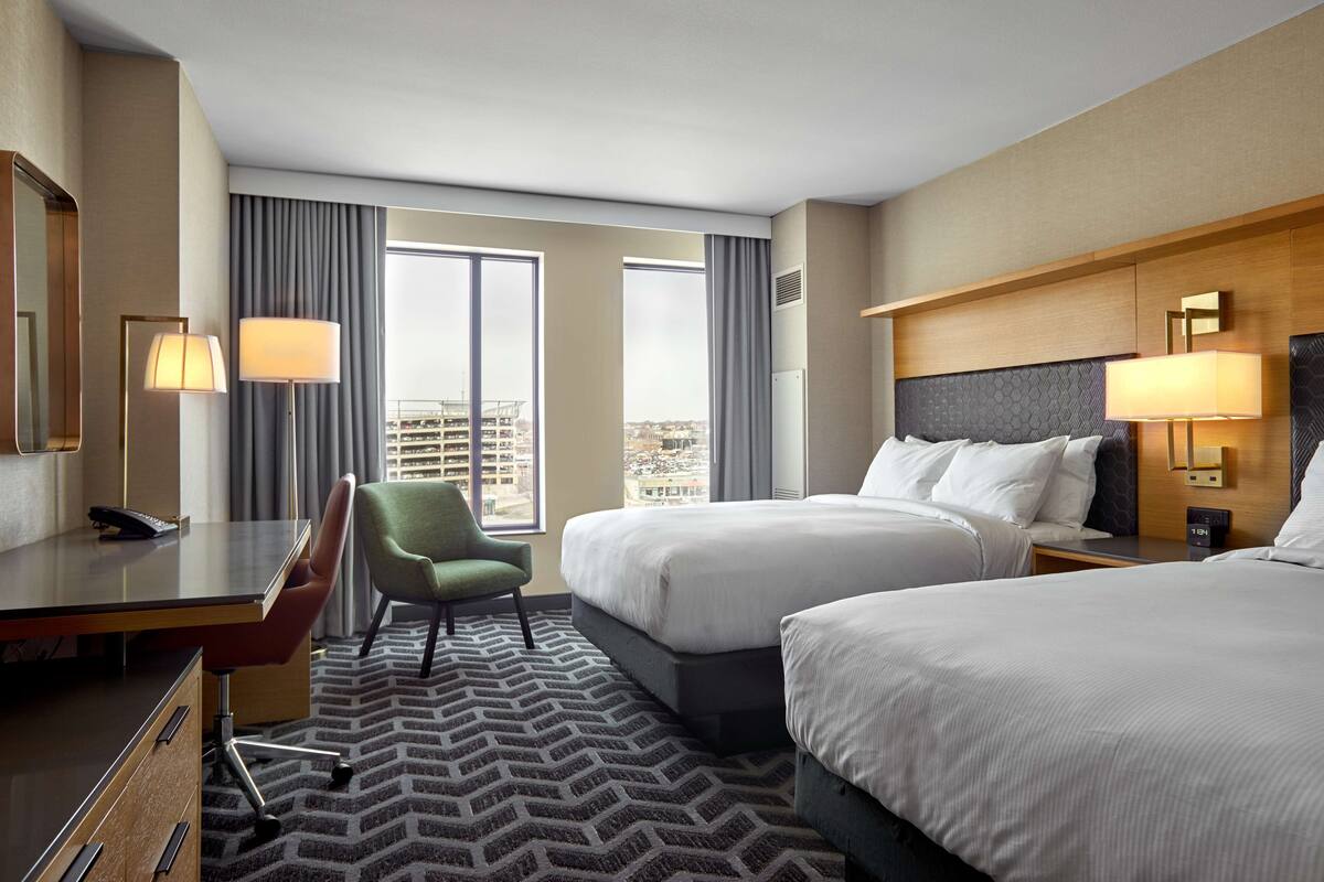 A hotel room with two beds, a desk, a chair, and a view of the city.