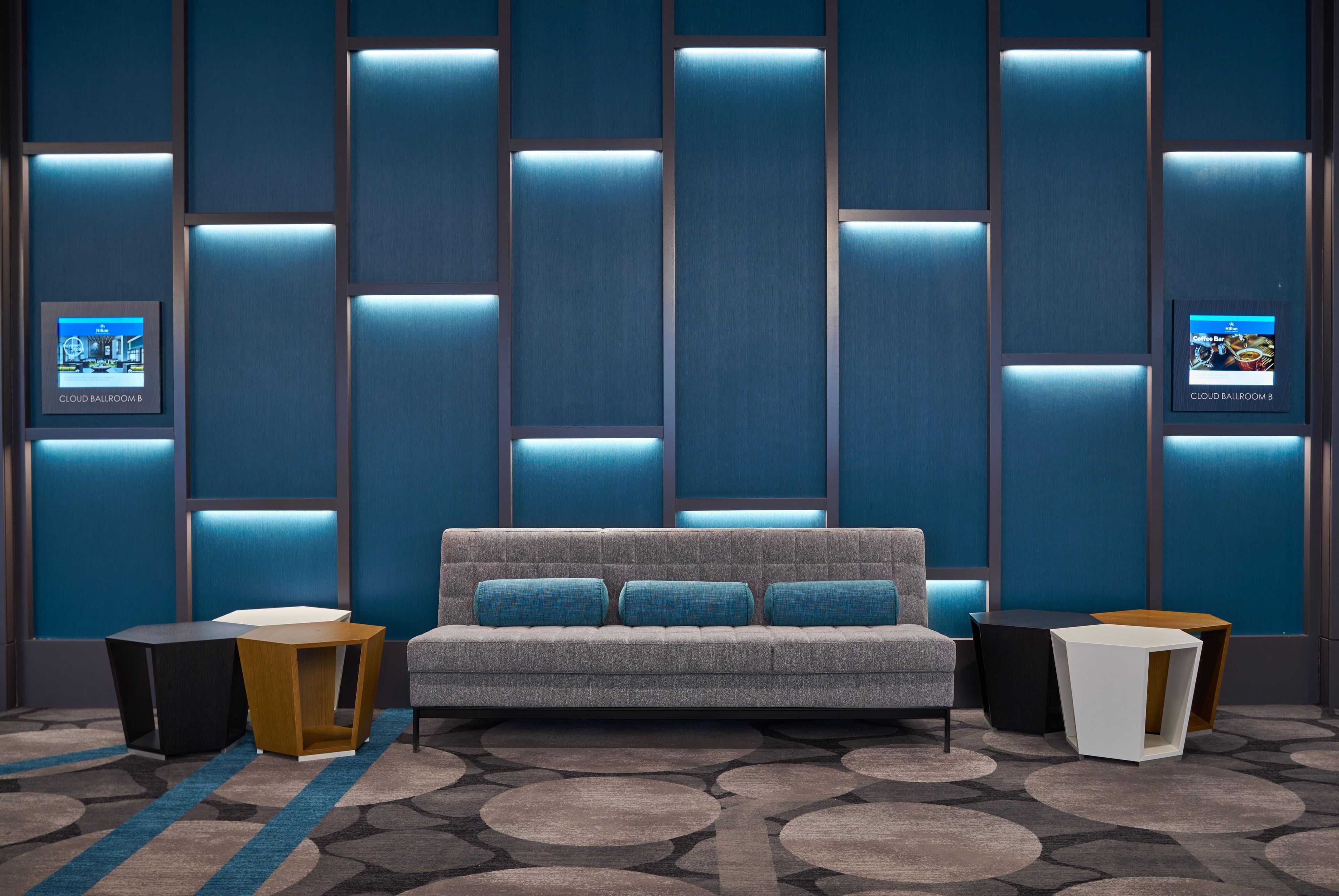 A modern hotel room with a grey sofa, two side tables, a patterned carpet, and a wall with illuminated panels.