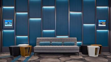 A modern hotel room with a grey sofa, two side tables, a patterned carpet, and a wall with illuminated panels.