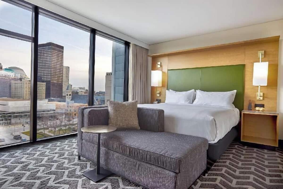 A hotel room with a large bed, a sofa, a small table, two bedside lamps, and a view of the city through large windows.