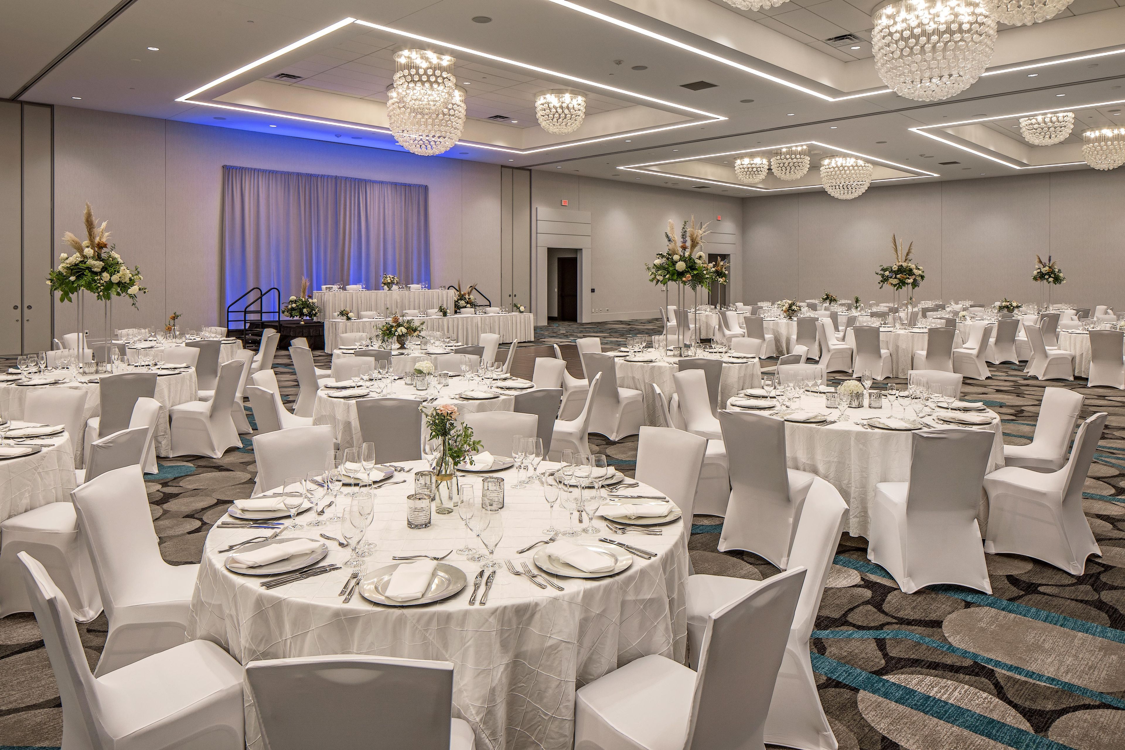 Une salle de banquet avec des tables rondes dressées pour le repas, des chaises blanches et des compositions florales décoratives.