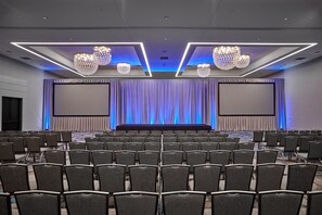 Un salón de conferencias con filas de sillas, un escenario y dos pantallas grandes.