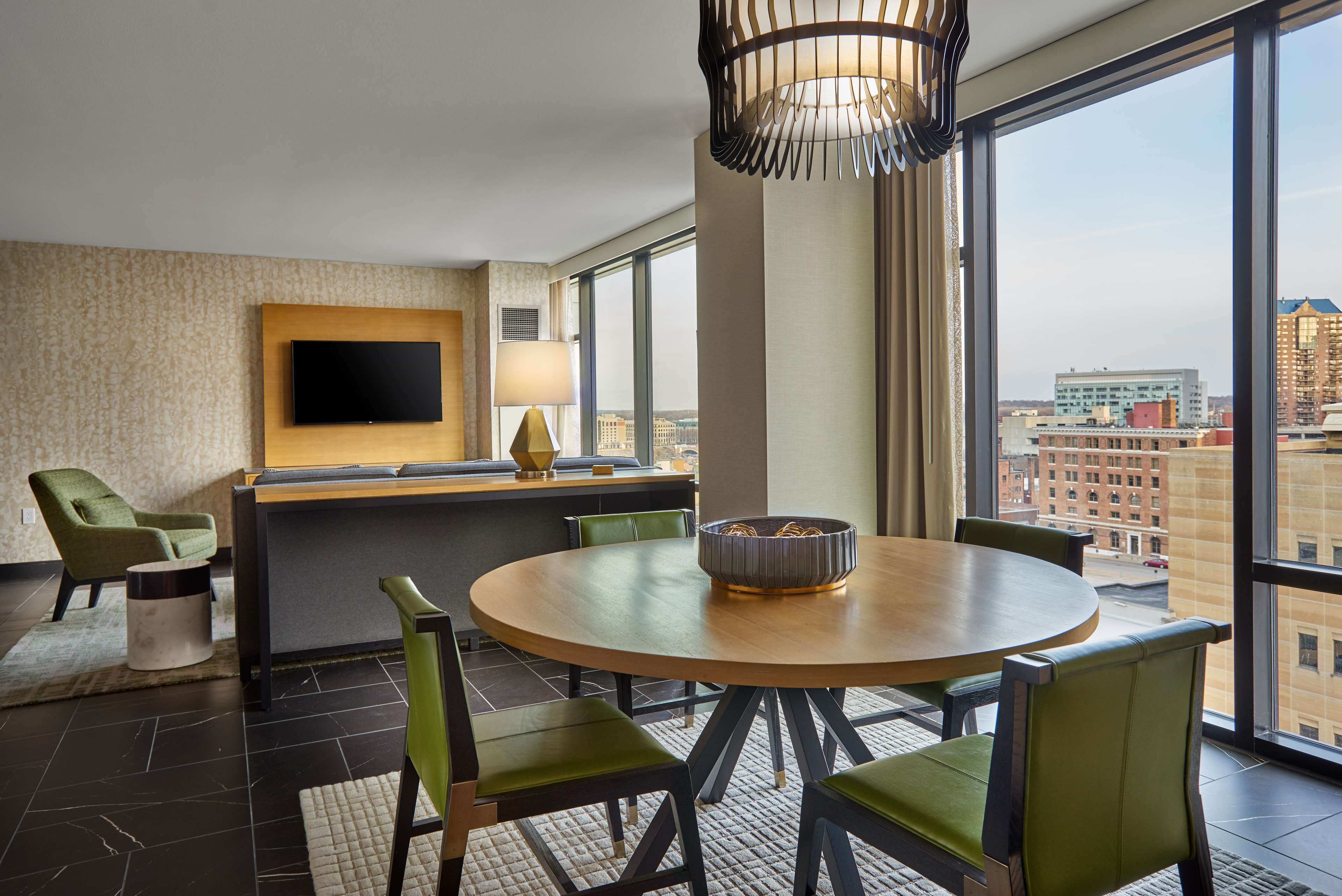 A modern hotel room with a round dining table, green upholstered chairs, a flat-screen TV mounted on the wall, and a large window offering a city view.