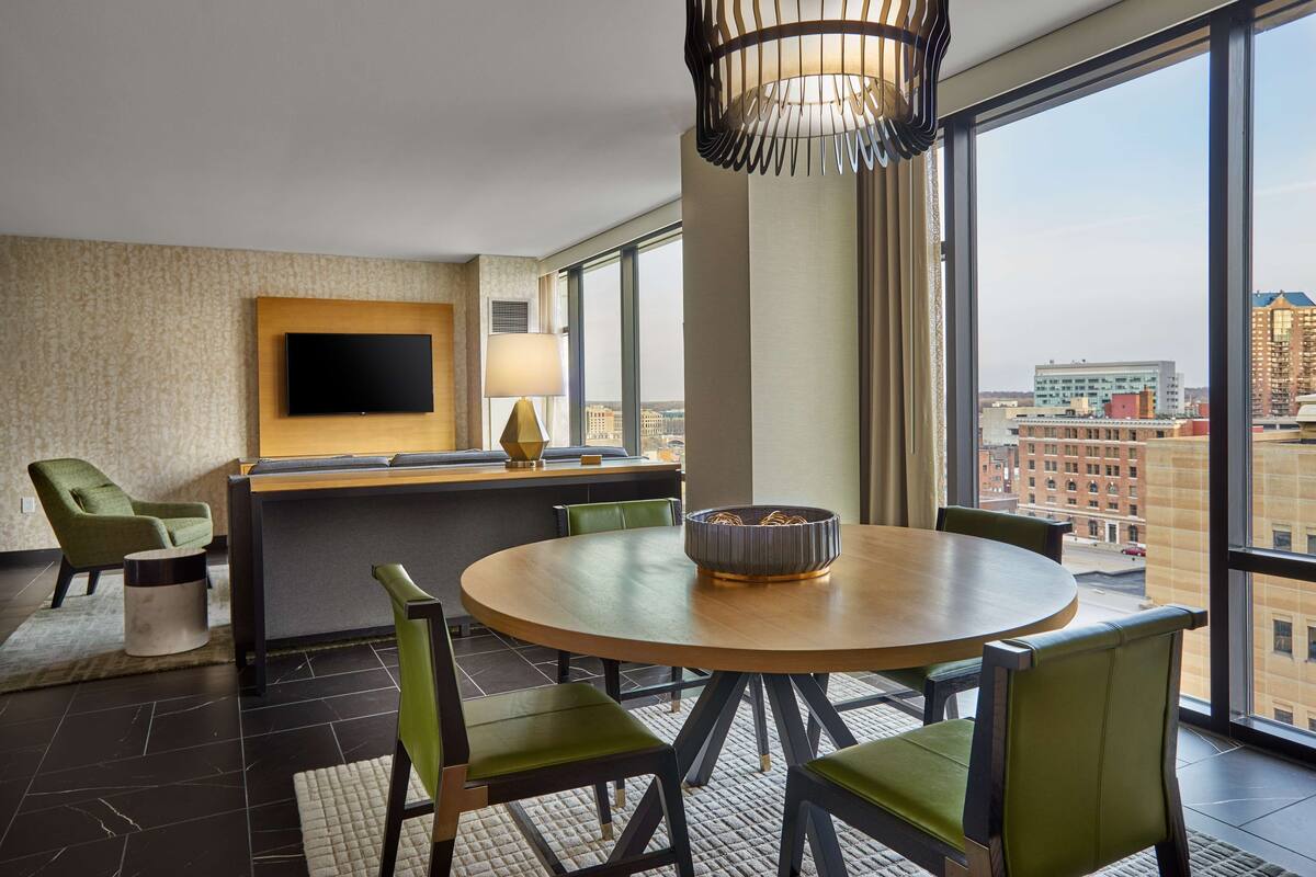A modern hotel room with a round dining table, green upholstered chairs, a flat-screen TV mounted on the wall, and a large window offering a city view.