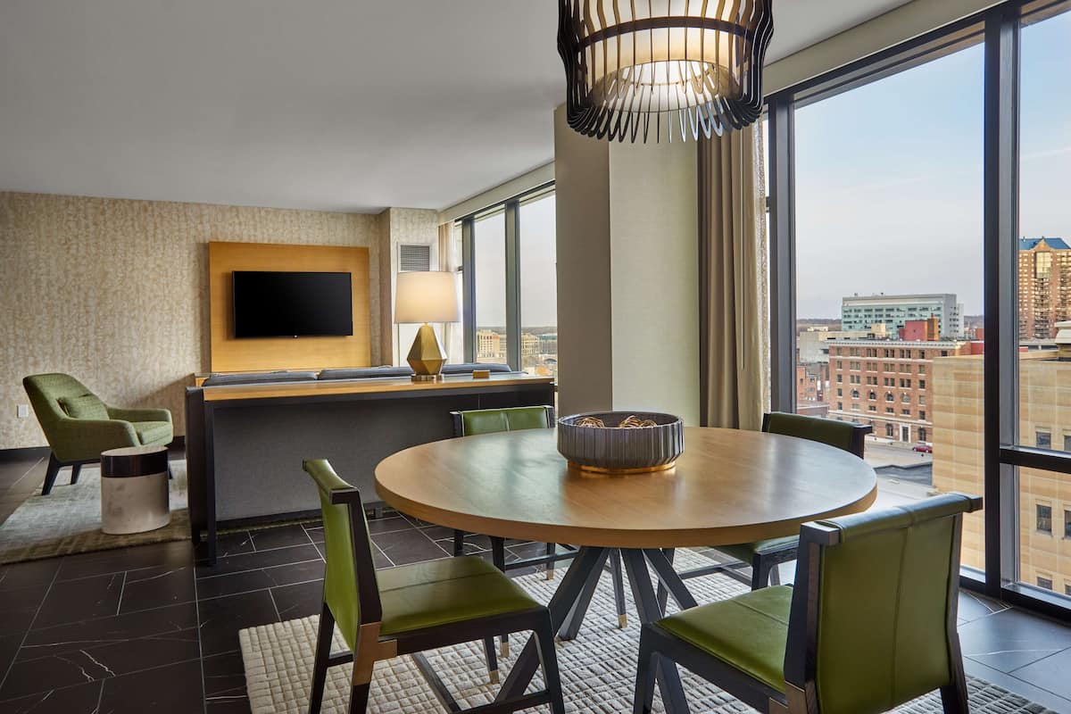 A modern hotel room with a round dining table, green upholstered chairs, a flat-screen TV mounted on the wall, and a large window offering a city view.