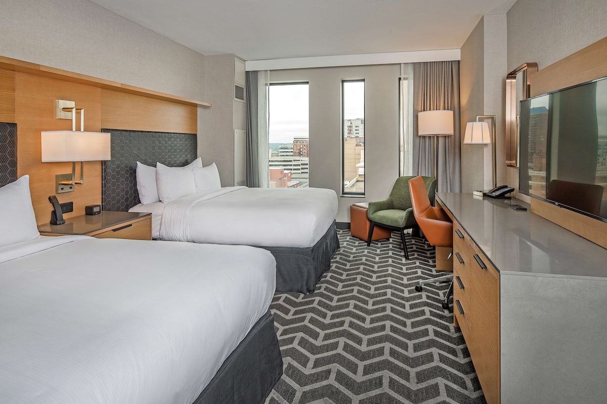 A hotel room with two beds, a desk, a chair, a TV, and a view of the city.