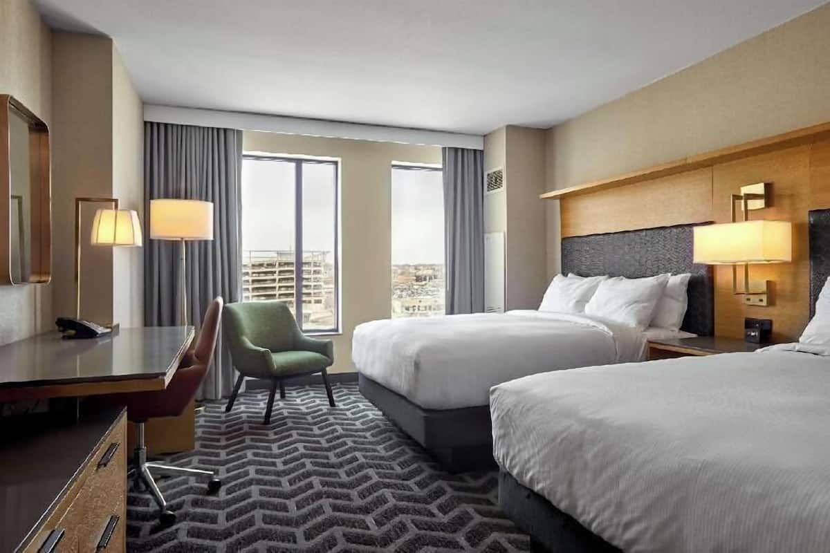 A hotel room with two beds, a desk, a chair, and a view of the cityscape.