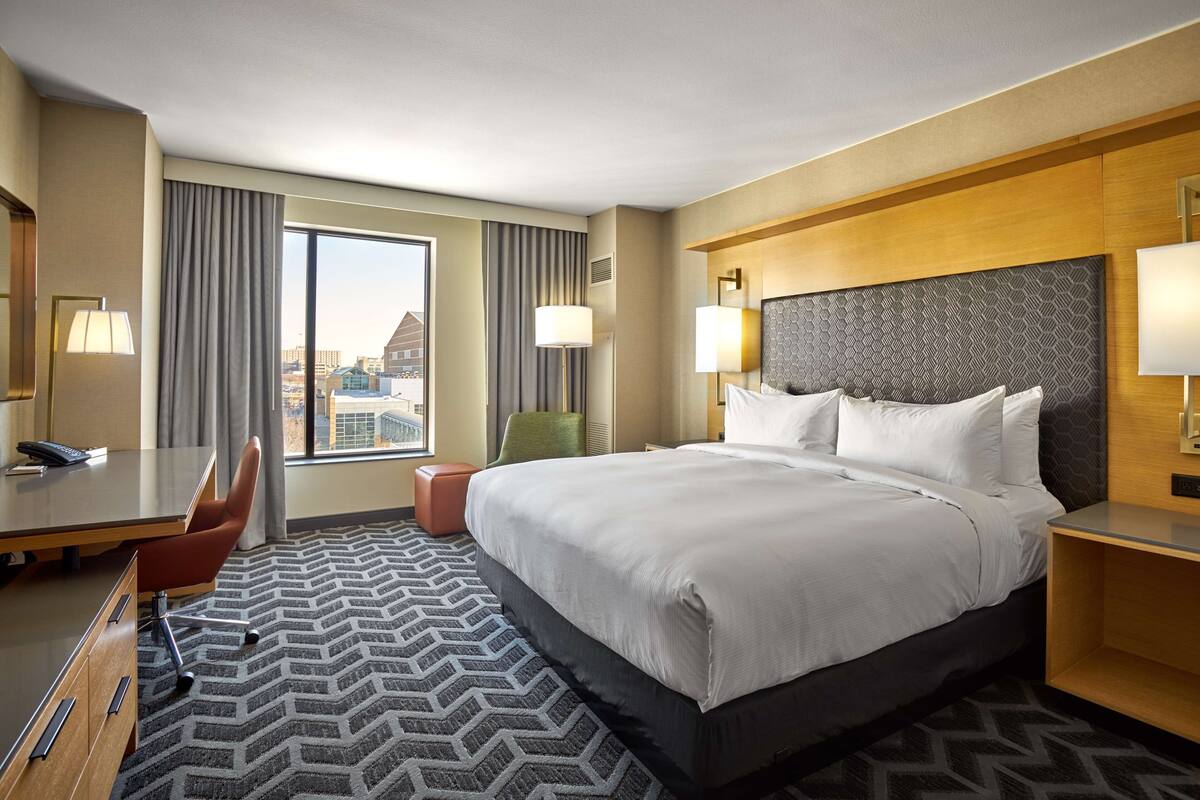 A hotel room with a large bed, a desk, a chair, and a view of the city.