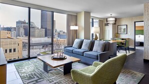 A modern hotel room with a large city view, a sofa, a dining area, and a small table with a decorative item.